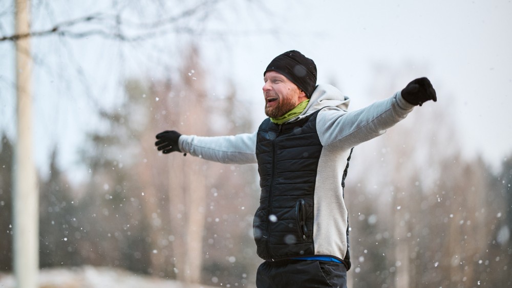 En man står i sportkläder och håller armarna utsträckta medan ett lätt snöfall faller omkring honom.